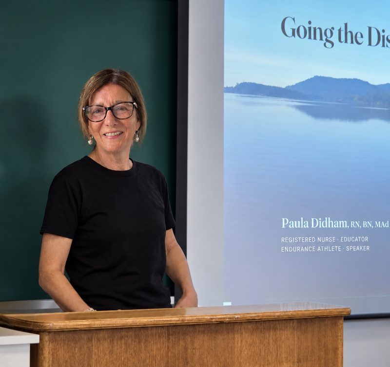 Paula speaking at a lectern in a classroom, her Going the Distance title slide projected on the screen beside her.