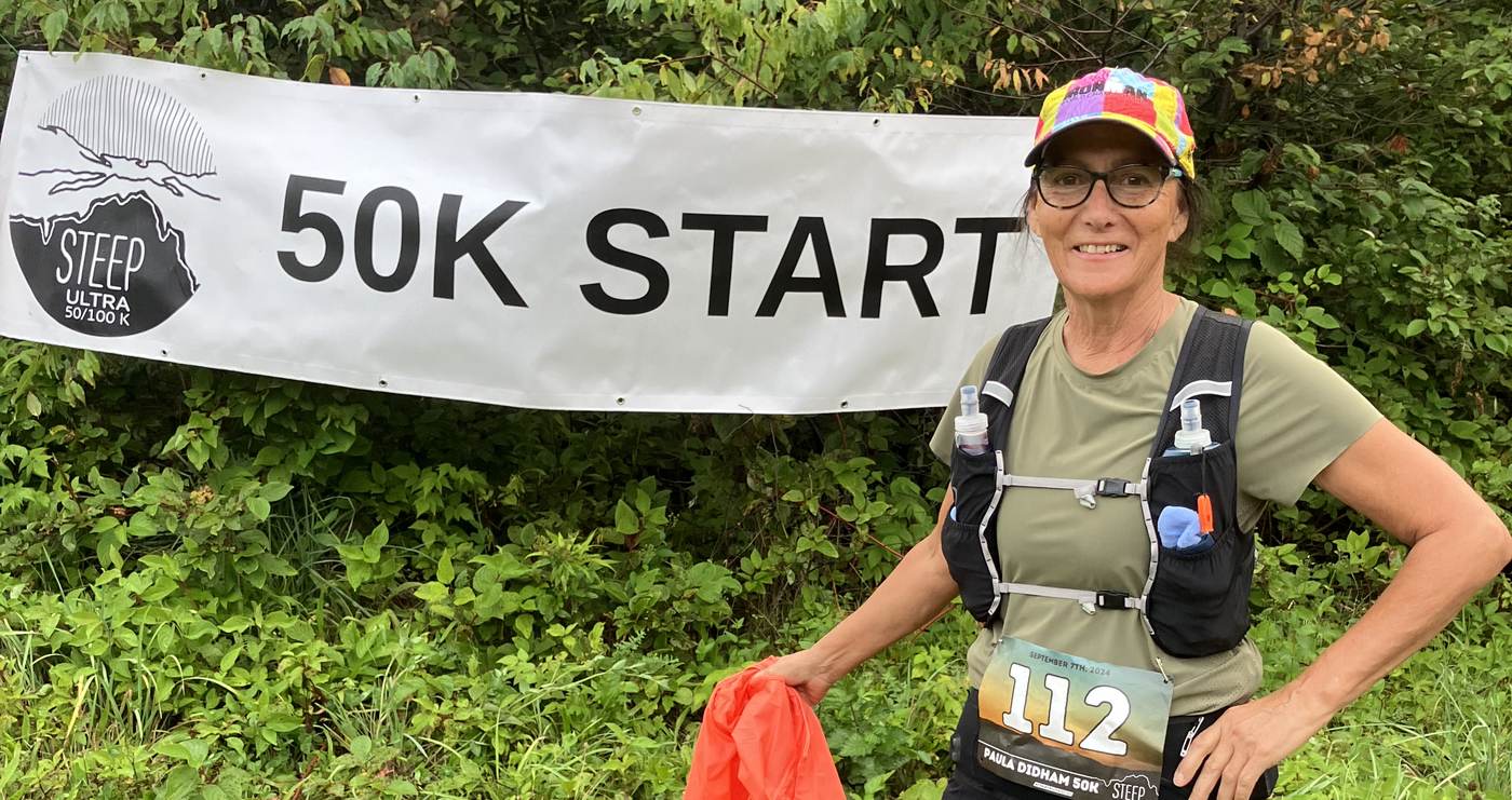 Paula at the 50K start banner of the STEEP Ultra trail race, smiling, wearing bib number 112 with her name on it.